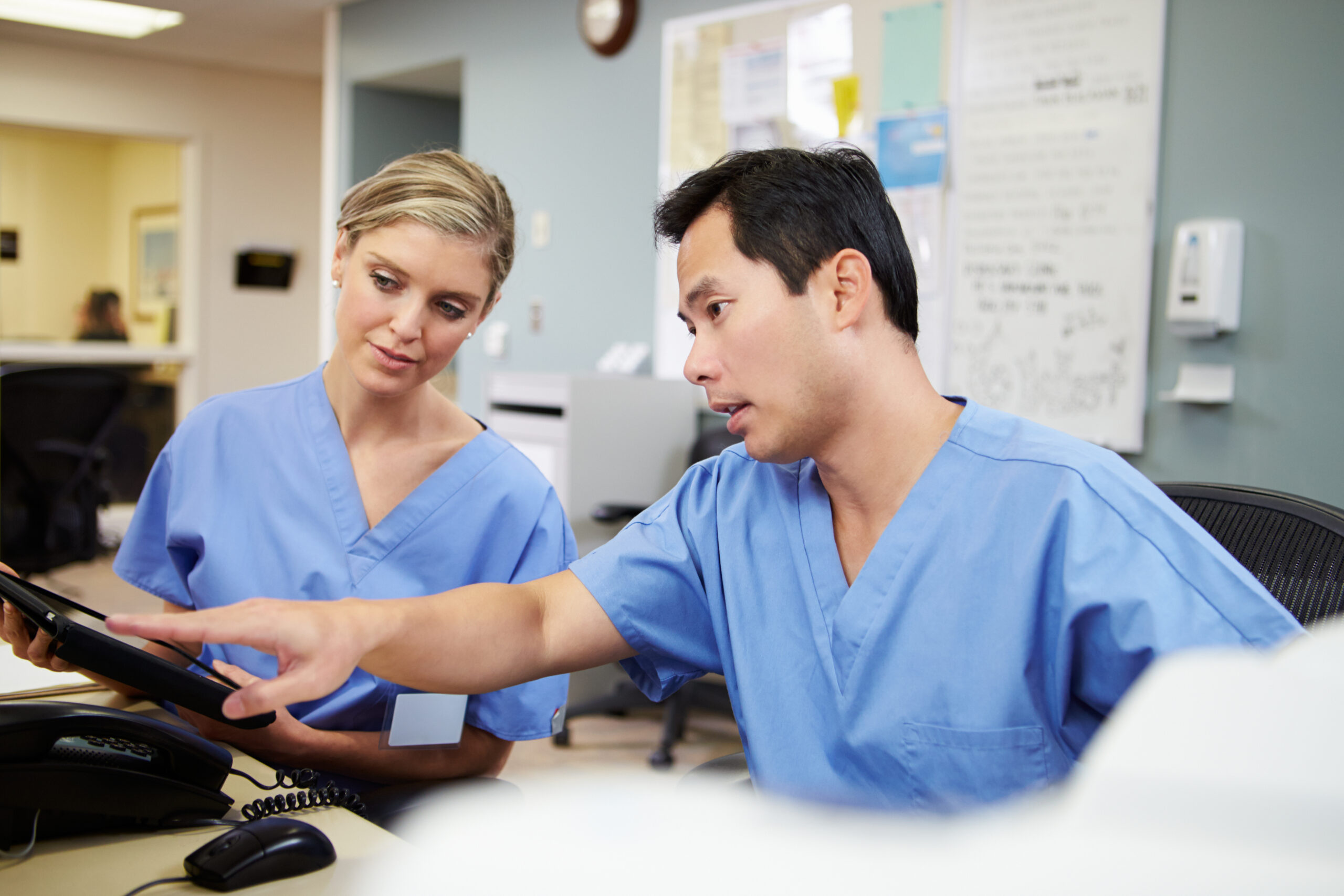 Two healthcare professionals in blue scrubs discuss information on a tablet, efficiently coordinating patient care with the aid of advanced nurse call systems in a clinical setting.