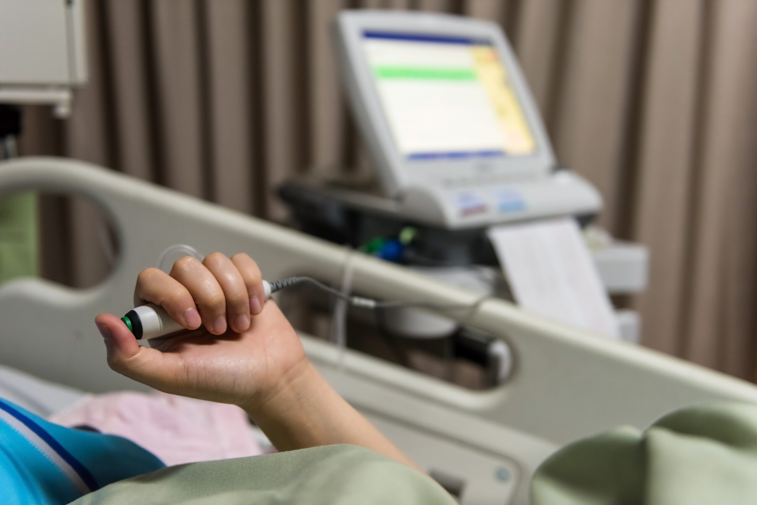 A person lies in a hospital bed holding a button connected to the advanced nurse call system, surrounded by medical equipment in the background.